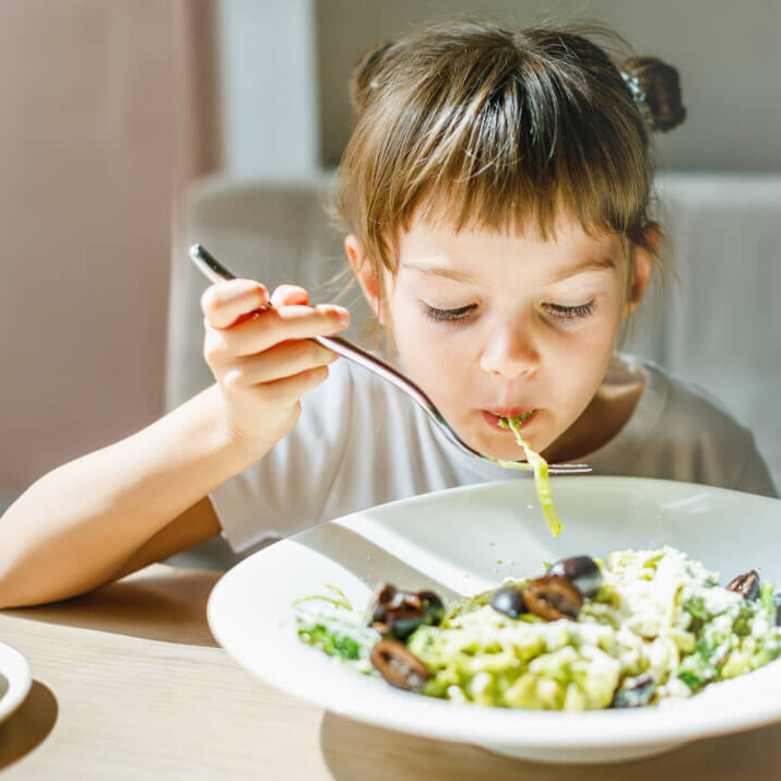 Young child happily eating a bowl of pesto pasta, with green sauce visible on the noodles, illustrating a creative way to incorporate more vegetables into meals. "Young child happily eating a bowl of pesto pasta, with green sauce visible on the noodles, illustrating a creative way to incorporate more vegetables into meals."