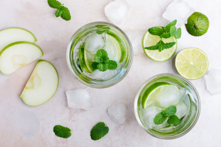 Two glasses of iced mint-lime beverages with lime wedges, apple slices, and fresh mint on a pale pink surface.