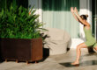 A woman in a green athletic outfit performs a high lunge yoga pose on a mat next to a large potted plant on a patio.