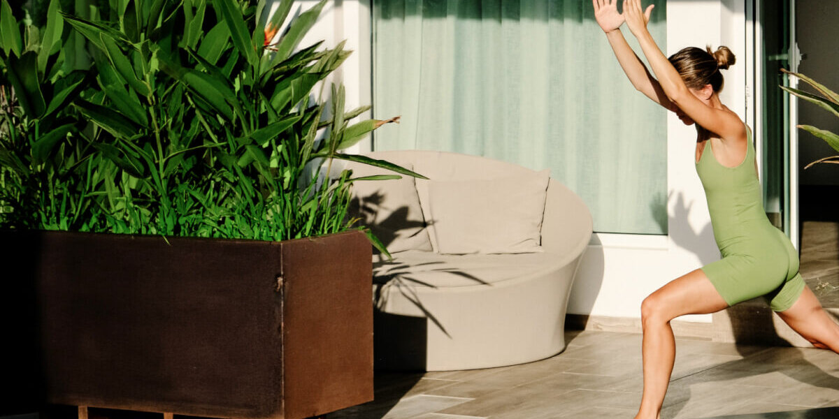 A woman in a green athletic outfit performs a high lunge yoga pose on a mat next to a large potted plant on a patio.