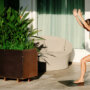 A woman in a green athletic outfit performs a high lunge yoga pose on a mat next to a large potted plant on a patio.