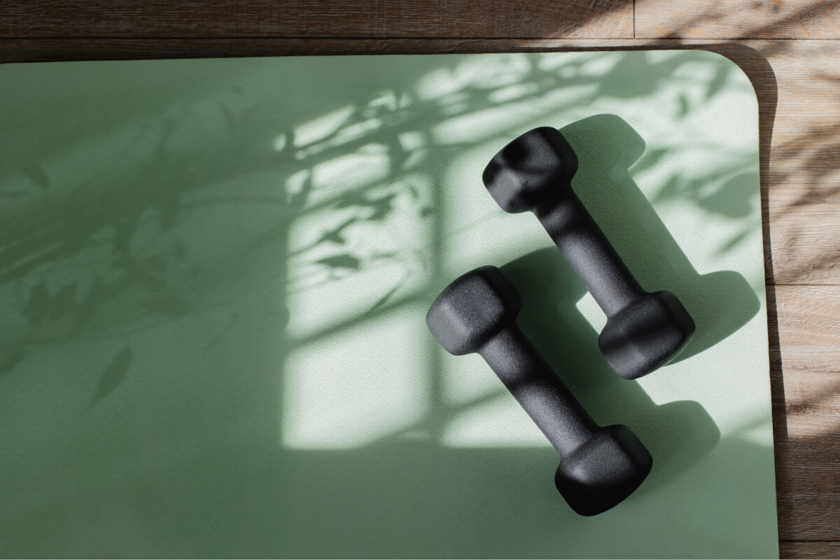 Two black dumbbells rest on a green exercise mat with shadowy leaf patterns.