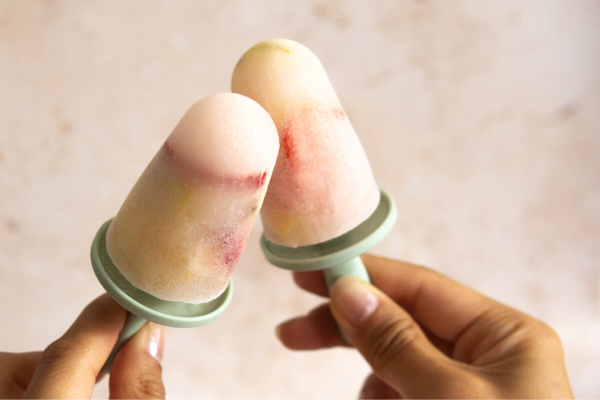 Two peach-colored popsicles on light green handles being held by a pair of hands.